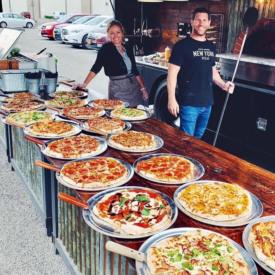 Two smiling chefs stand behind a long table laden with various pizzas, next to a food truck.