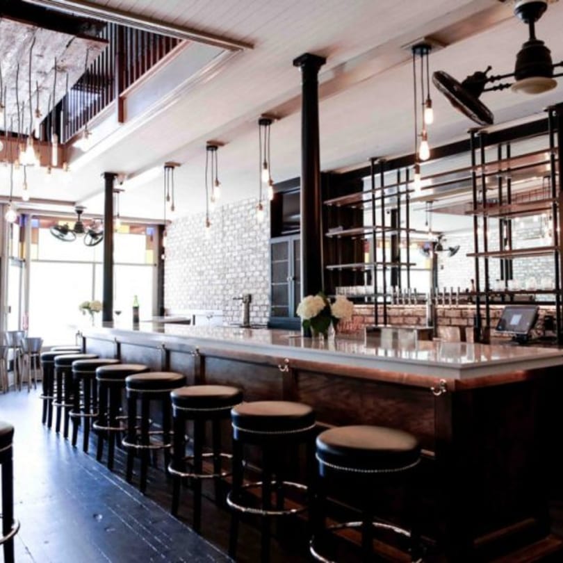 A long, dark wooden bar with black stools, exposed brick, and industrial lighting in a restaurant.
