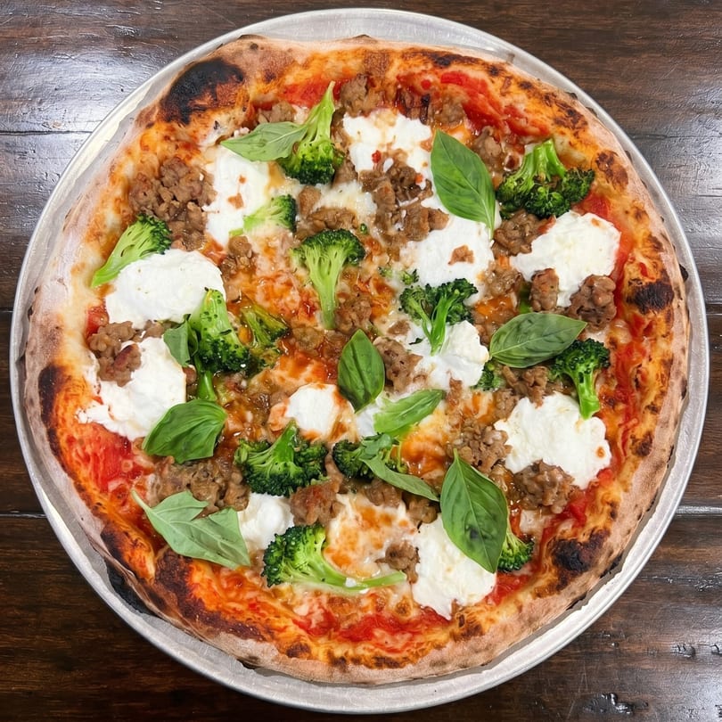 A whole pizza with sausage, broccoli, fresh mozzarella, and basil on a metal pan.