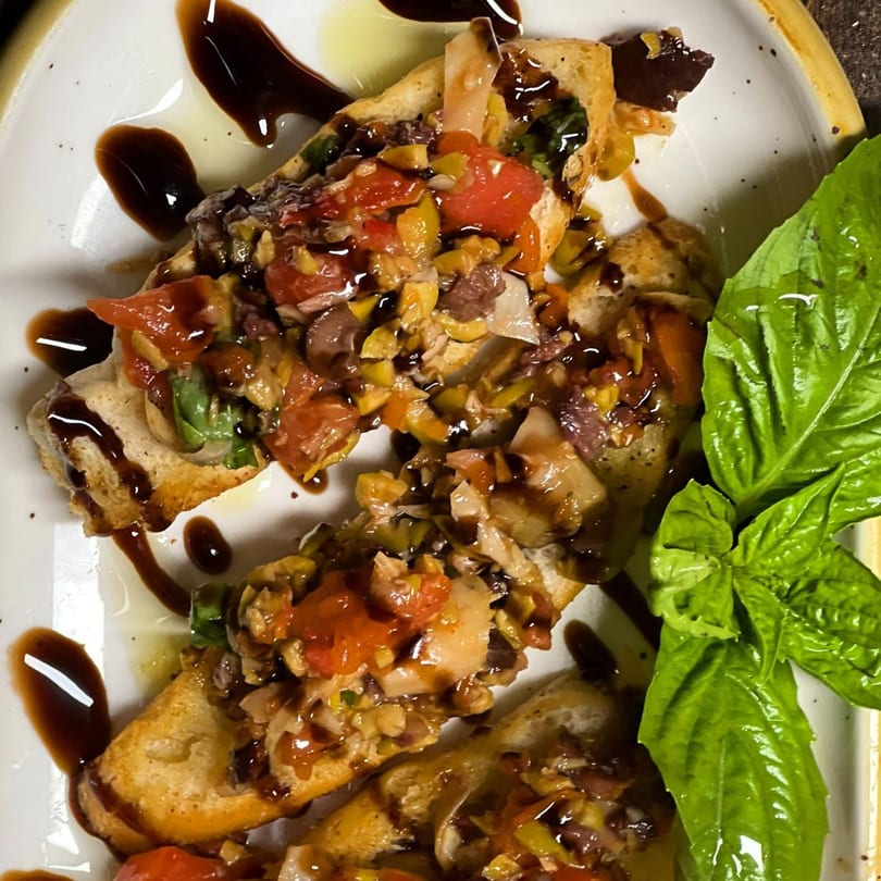 Bruschetta topped with diced tomatoes, olives, and herbs, drizzled with balsamic glaze and olive oil, garnished with fresh basil.