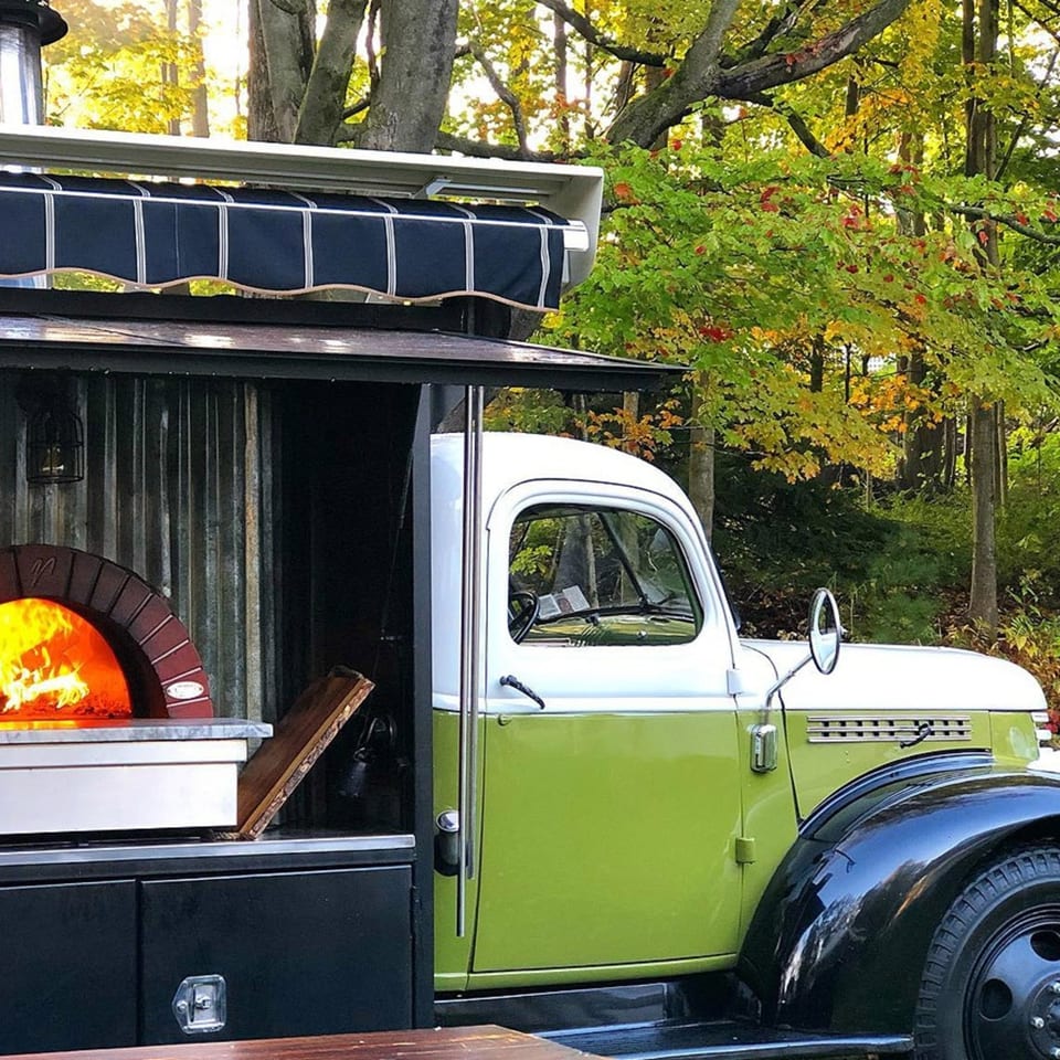 A vintage green and white food truck with a wood-fired pizza oven, set against autumn trees.