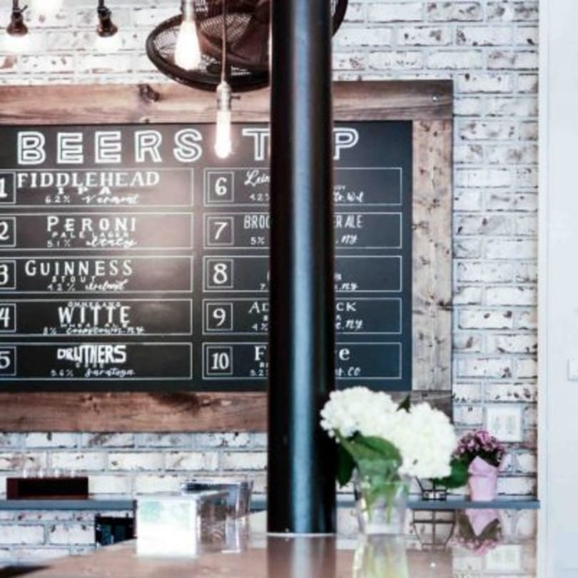 A chalkboard beer menu with various brews listed, set against a white brick wall.