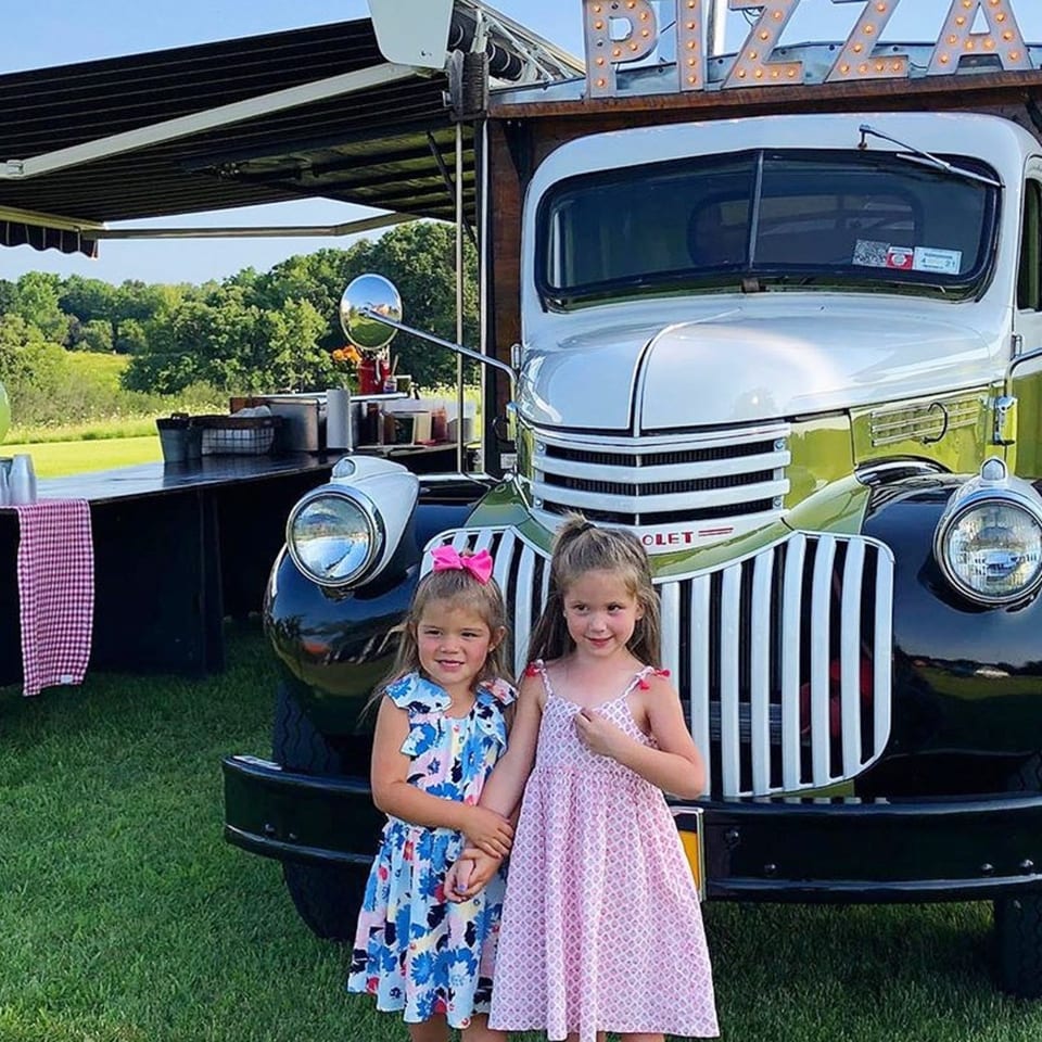 Two young girls stand in front of a vintage pizza food truck on a grassy field.