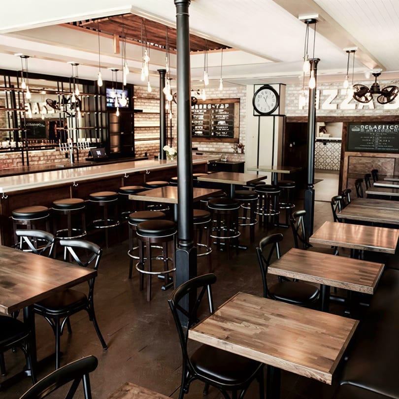 Interior of a restaurant with wooden tables, black chairs, bar stools, and brick walls.